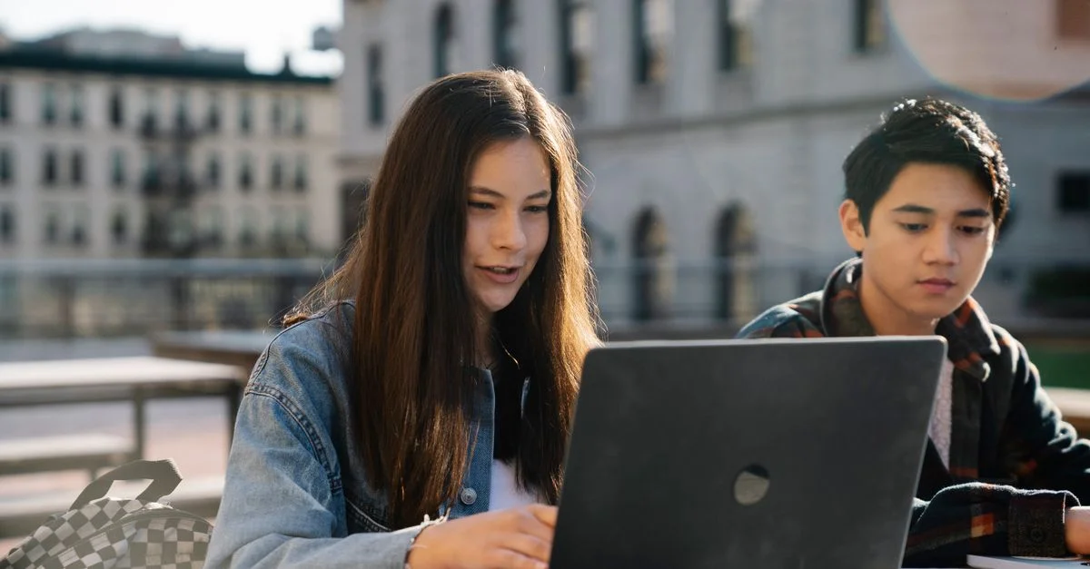 Deux étudiants collaborant sur un ordinateur portable en extérieur avec un bâtiment historique en arrière-plan.