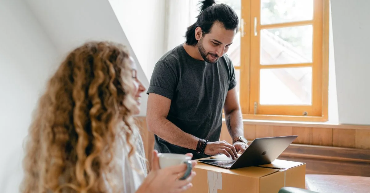 Couple heureux déballant dans leur nouvelle maison, utilisant un ordinateur portable sur une boîte, partageant du café et des sourires.