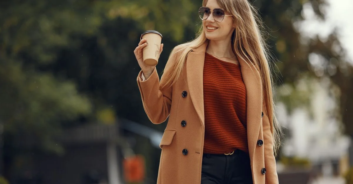 Femme élégante en manteau camel dégustant un café par une journée ensoleillée en extérieur.