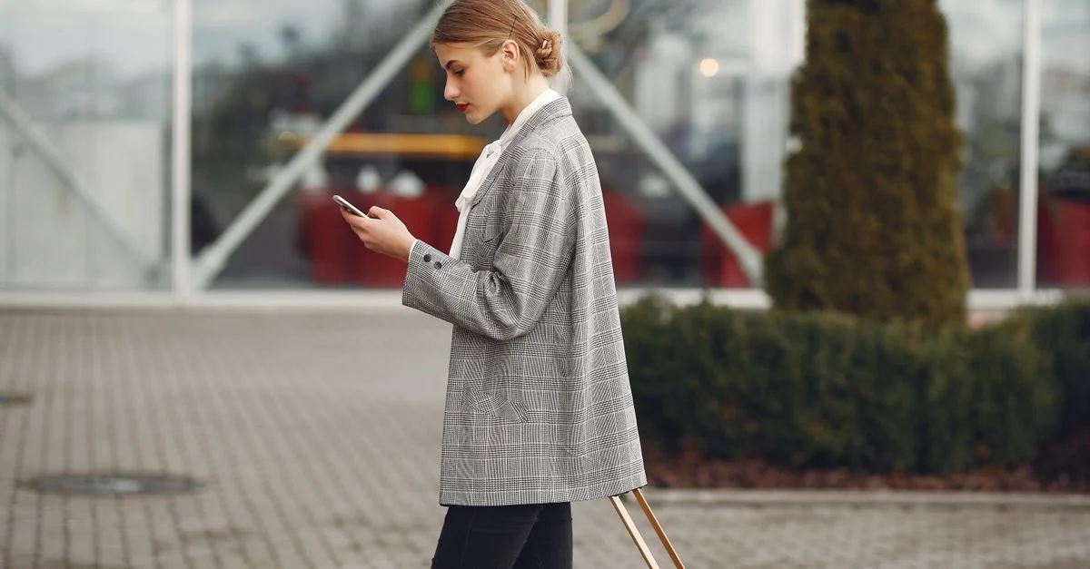 Femme élégante marchant avec une valise tout en utilisant son smartphone devant un bâtiment moderne.